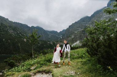 Boho elbisesi içinde güzel bir gelin, bir şapka ve bir damat dağlarda gülüyor ve gülüyor. Gelin ve damat dağlarda sarılıp yürür.