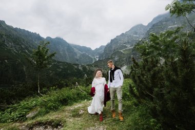 Boho elbisesi içinde güzel bir gelin, bir şapka ve bir damat dağlarda gülüyor ve gülüyor. Gelin ve damat dağlarda sarılıp yürür.