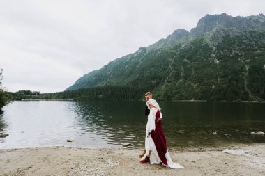 Damadın ve boho elbisenin içindeki güzel gelin dağlarda bir gölün kenarında gülüyor ve gülümsüyor. Gelin ve damat göl kenarındaki dağlarda sarılıp yürüyorlar..