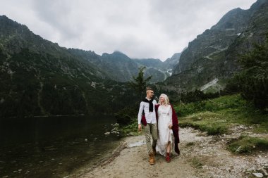 Damadın ve boho elbisenin içindeki güzel gelin dağlarda bir gölün kenarında gülüyor ve gülümsüyor. Gelin ve damat göl kenarındaki dağlarda sarılıp yürüyorlar..