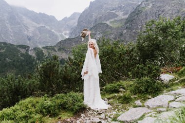 Boho elbisesi içinde güzel bir gelin elinde bir şapkayla gölün kenarındaki dağlarda gülümsüyor. Güzel gelin dağlarda gülümser ve güler..