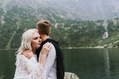 Damadın ve boho elbisenin içindeki güzel gelin dağlarda bir gölün kenarında gülüyor ve gülümsüyor. Gelin ve damat dağlarda kucaklaşıyorlar..