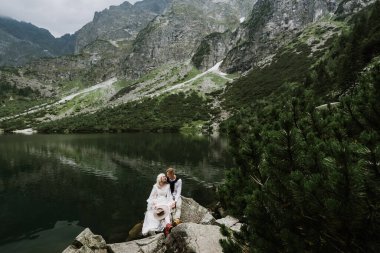 Boho elbisesi içinde güzel bir gelin ve damat dağlardaki bir gölün yanındaki taşların üzerinde oturuyorlar. Gelin ve damat dağlarda bir göl kenarında gülüyor ve gülüyorlar.
