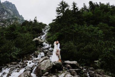 Boho tarzı bir elbise içinde güzel bir gelin, şapka ve damat dağlarda küçük bir şelalenin yanında sarılıp öpüşüyor..