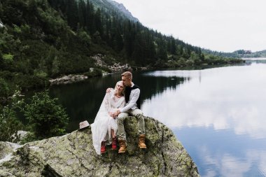 Boho tarzı bir elbise içinde güzel bir gelin. Şapka ve damat dağlardaki bir gölün kenarında taşların üzerinde oturuyor..