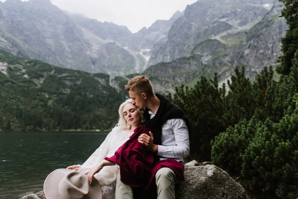 美丽的新娘穿着紧身衣和新郎 在山中的一个湖边笑着 新娘和新郎在湖边的山上拥抱 — 图库照片