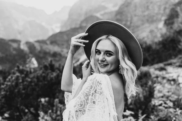 Beautiful bride in a dress and hat and smiling in the mountains by the lake. Beautiful bride smiles and laughs in the mountains. Black and white photo of a bride in the mountains.