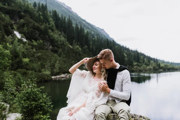 美丽的新娘穿着布荷族风格的衣服 帽子和新郎坐在高山湖畔的石头上 — 图库照片
