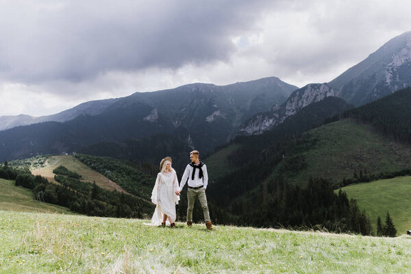 Beautiful bride in a boho style dress, hat and groom walk and laugh in the mountains.