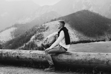 Bir adam dağlarda bir kütüğe oturur. Dağlarda bir kütüğün üzerindeki siyah-beyaz bir adamın fotoğrafı..