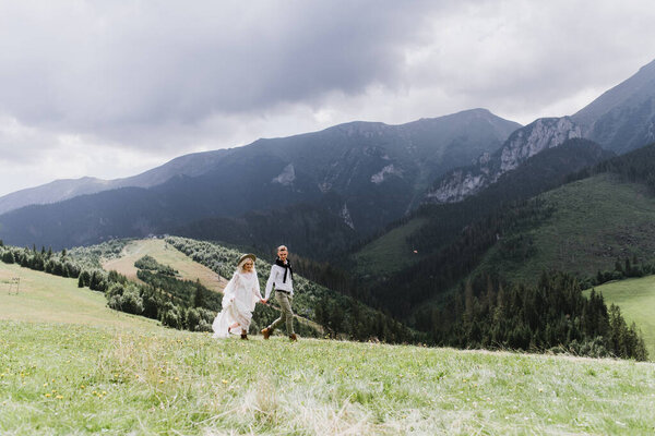 Beautiful bride in boho style dress, hat and groom run and laugh in the mountains.