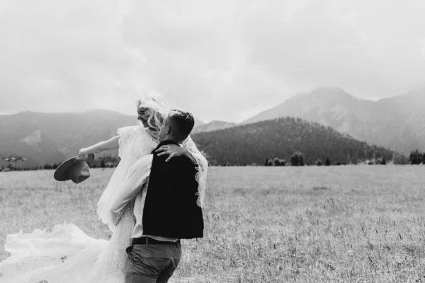 The groom circles the bride in his arms in the mountains. The groom holds the bride in a boho dress and a hat in his hands in the mountains. The bride waving her hat in the mountains.