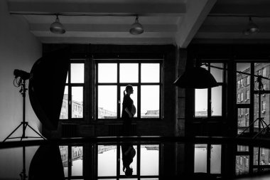 Pencerenin yanında siyah takım elbiseli hamile bir kadın var. Stüdyoda pencerenin önündeki hamile bir kadının siyah beyaz fotoğrafı..