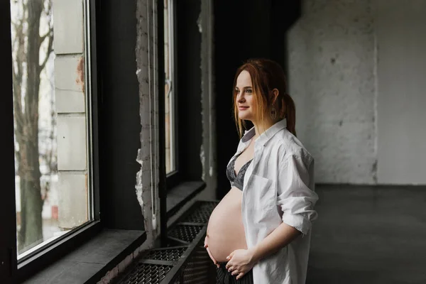 穿着内裤和白衬衫的年轻孕妇站在演播室窗前 — 图库照片