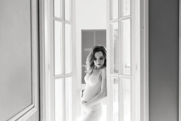 年轻的孕妇穿着白色的紧身衣在玻璃门附近的工作室里 在演播室门口 一张怀孕妇女的黑白照片 — 图库照片