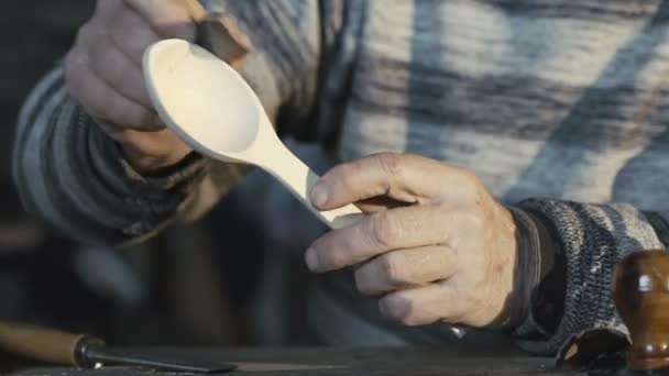 Old wrinkled carpenter hews wooden spoon and smiling at camera — Stock Video