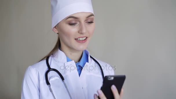 Νεαρή γυναίκα γιατρό κάθεται με έξυπνο κινητό τηλέφωνο στο νοσοκομείο και χαμογελαστός — Αρχείο Βίντεο