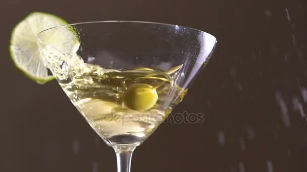 Оливка падает в бокал со спиртом и ломтиком лайма. Медленно. — стоковое видео