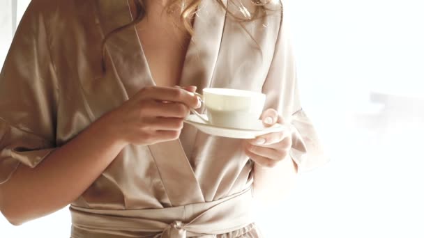 Uma rapariga bonita à noite bebe uma chávena de café perto da janela. Devagar. — Vídeo de Stock