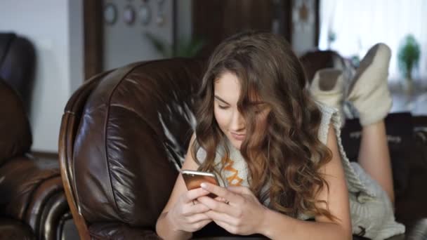 Chica bonita relajarse en el sofá de cuero y el uso de teléfono móvil 4K — Vídeo de stock