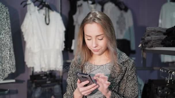 Chica bonita usando el teléfono móvil en una tienda de ropa. 4K — Vídeos de Stock