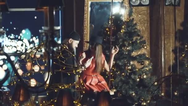 Man pushes apart girl on a swing indoor with trees and garlands — Stock Video