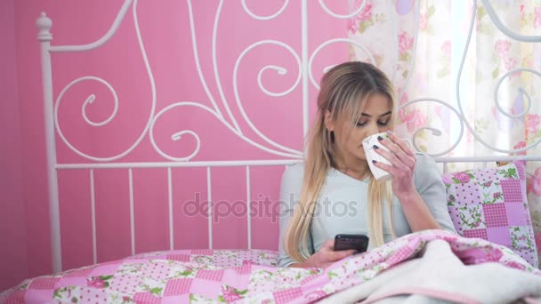 Pen blondine i senga, som drikker og bruker mobiltelefon i 4K. – stockvideo