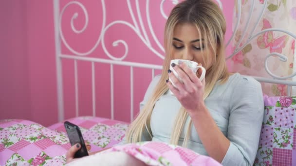 Mooie blonde liggend in bed, drinken en het gebruik van mobiele telefoon in 4k — Stockvideo