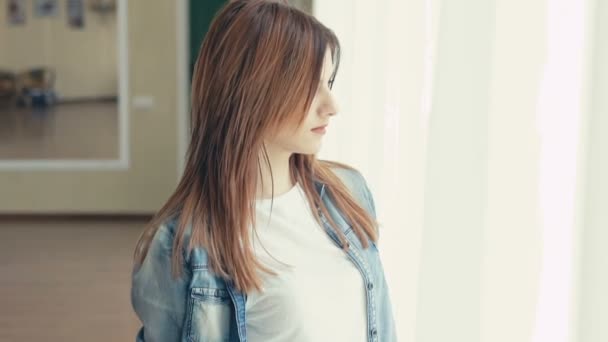 Porträt eines hübschen Mädchens, das auf dem Fensterhintergrund in die Kamera blickt. langsam — Stockvideo