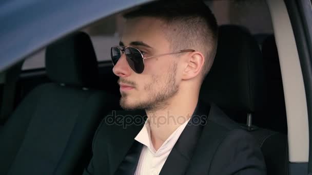 Молодой человек в солнцезащитных очках смотрит и улыбается из окна автомобиля 4K — стоковое видео