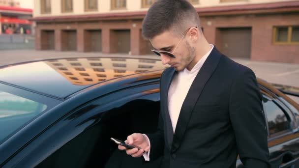 Joven hombre de negocios guapo utilizando el teléfono móvil cerca del coche de lujo 4K — Vídeo de stock