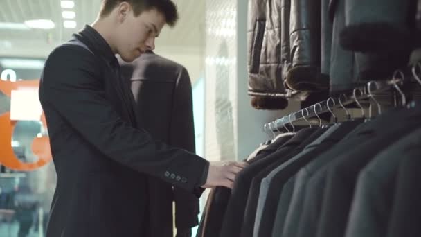 Элегантный молодой человек в костюме выбирает одежду в магазине одежды — стоковое видео