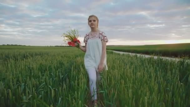 Молода мила дівчина у вишитій сукні гуляє на пшениці з букетом квітів — стокове відео