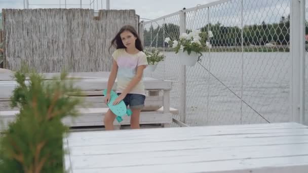 Underbara lilla flicka på en strandpromenad poserar, hålla hennes skateboard — Stockvideo