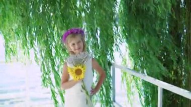 Küçük sarışın çiçek taç ve ellerinde çiçeklerle kameraya 4 k gülümsüyor