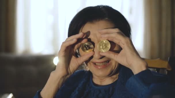 Portrait of old woman puts on two golden bitcoins to glasses and smiles — Stock Video