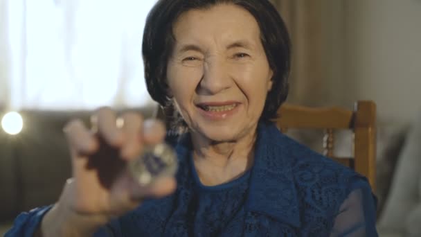 Portret van oude lachende vrouw houdt een zilveren bitcoin in de hand op camera — Stockvideo