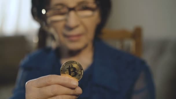 Portrait of old woman holds a golden bitcoin in hand at camera — Stock Video