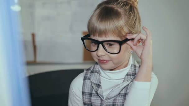 Симпатичная маленькая улыбчивая девушка в очках охотно работает с компьютером в офисе. 4K — стоковое видео