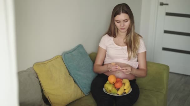 Ευτυχισμένη έγκυος γυναίκα ξεφλούδισμα κρούστα μανταρίνια στον καναπέ στο σπίτι — Αρχείο Βίντεο