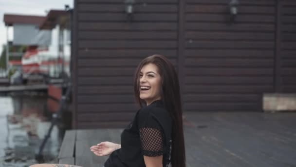 Happy brunette relaxing and waving legs on the deck at lake, smiling and posing — Stock Video