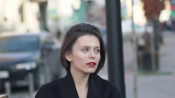 Stijlvolle dame in zwarte jas en rode lippen poseert op camera op winderige straat — Stockvideo