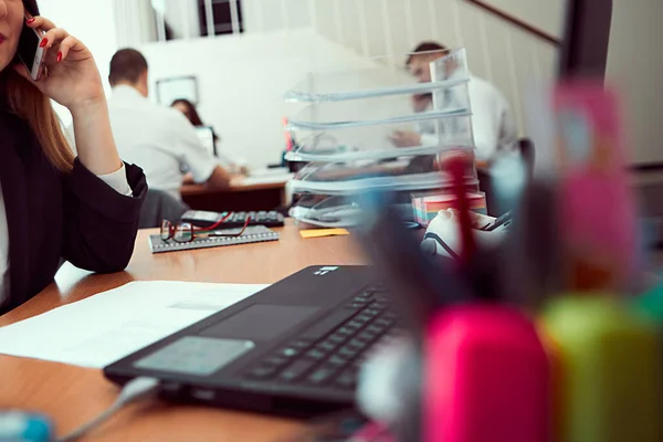 Young business woman working in a new modern office at a table on a laptop and talking on a cell phone. In the background are a group of employees working. IT sphere of life concept.