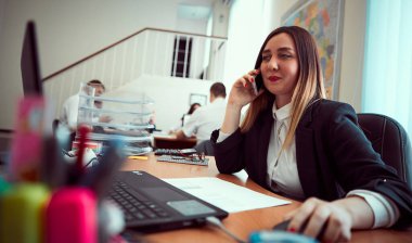 Modern bir ofisin bilgisayar masasında çalışmak için dizüstü bilgisayar kullanan mutlu, neşeli, güzel bir iş kızının yakın plan portresi..