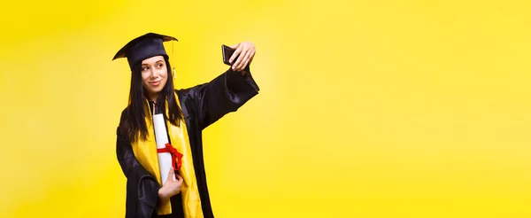 graduate woman makes selfie photo on phone over yellow background, concept of successful completion of studies, panoramic mock-up with place for text