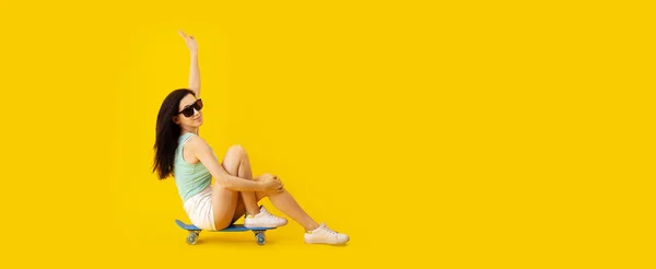 照片上年轻姑娘戴着太阳镜坐在滑板上 背景黄色 — 图库照片