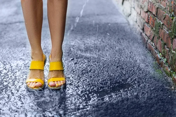 Female feet standing in the rain on wet pavement near a brick wa