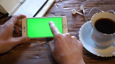 adam evde onun smartphone üzerinde slaytlar. yeşil ekran. Mobil aygıtın yanında her iki ahşap bir masa üzerinde kahve fincanı.