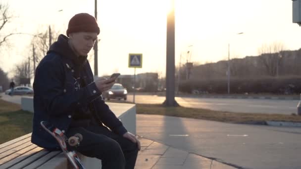 Homem sentado em seu skate e mensagens de texto, pôr do sol fundo . — Vídeo de Stock