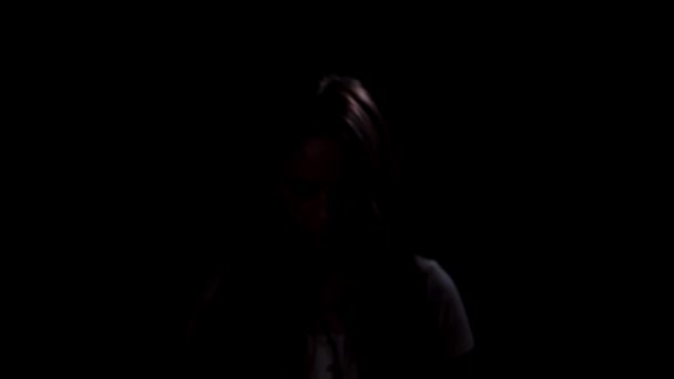 A tearful and frightened girl comes out of the darkness and looks in the camera on a black background. — Stockvideo